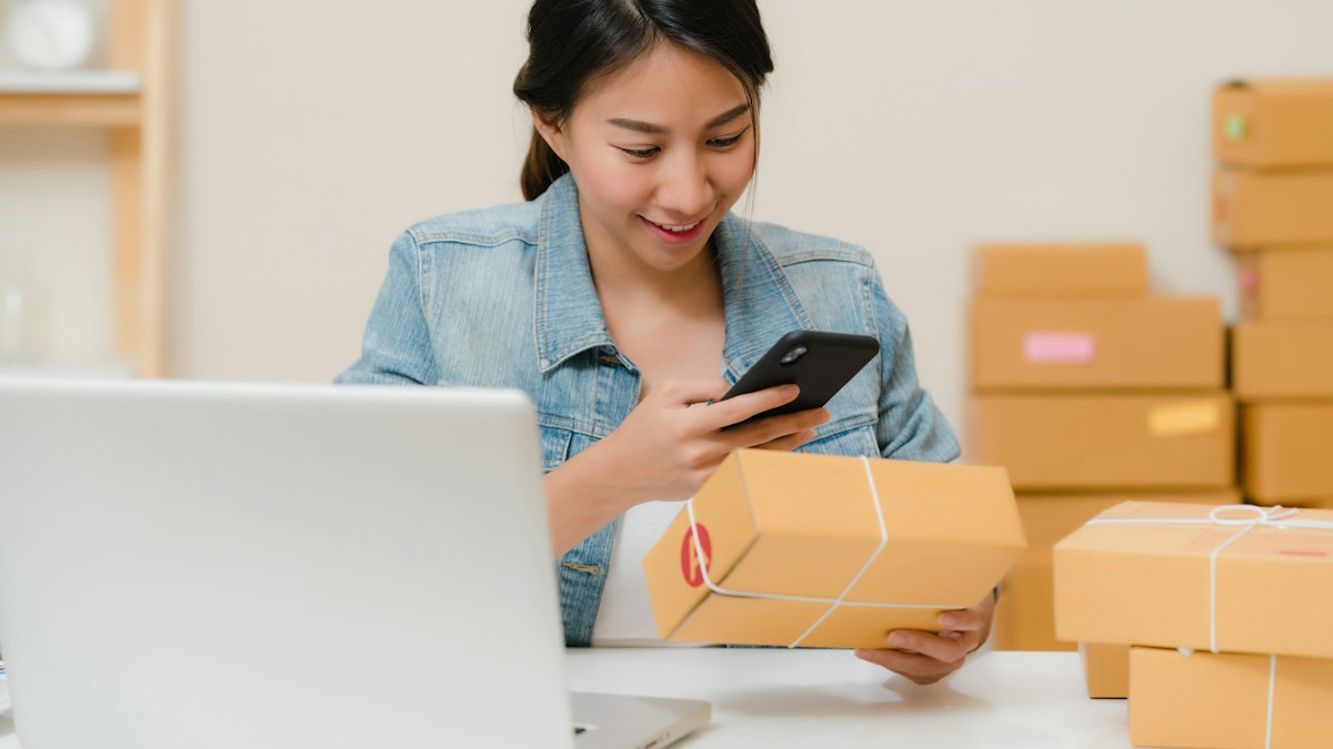 A seller checking an order on her phone next to parcels and a laptop